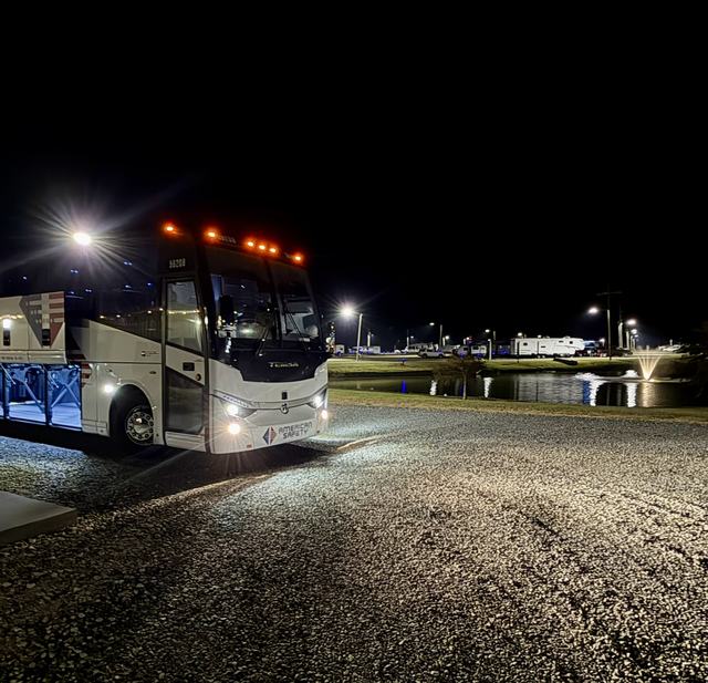 White charter bus with headlights on parked on gravel at night near a pond