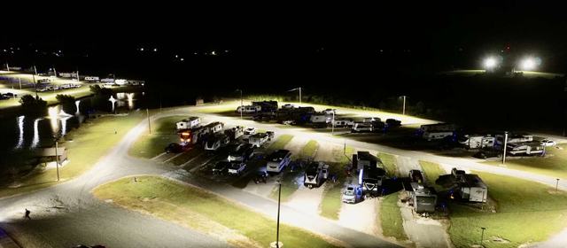Night aerial view of lit RV sites with parked RVs and vehicles near a pond