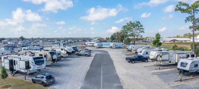 Paved road running between gravel RV sites with trailers and trucks toward a pond