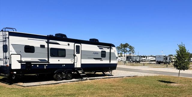 Travel trailer parked on a gravel site beside grass and a young tree